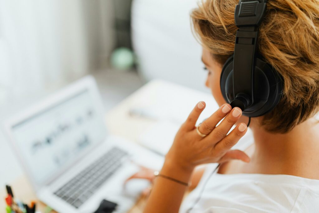 A woman with short light brown hair wearing black headphones while seated in front of a white notebook computer; she touches one hand to one of the ear cups.