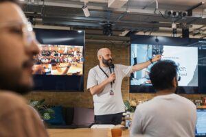 An example of how to make your content more engaging: A man with a beard wearing a white t-shirt and lanyard presents to a group in what appears to be a casual office space. He's pointing a hand to his left as people in the foreground look on.